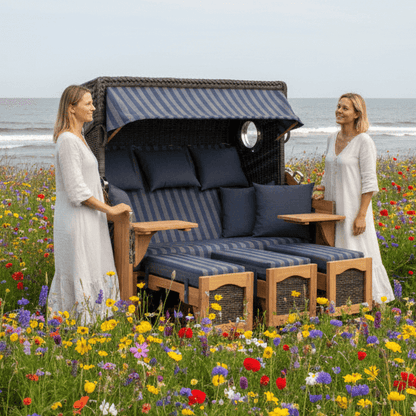Strandkorb deVries PURE S Seemöwe Teak mit Bullaugen und blauen Polstern im Blumenfeld am Meer