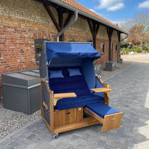 Strandkorb Prieß, Modell Meister mit Iroko Holz und dunklem PE-Geflecht, blau gestreift, in einem Garten setting.