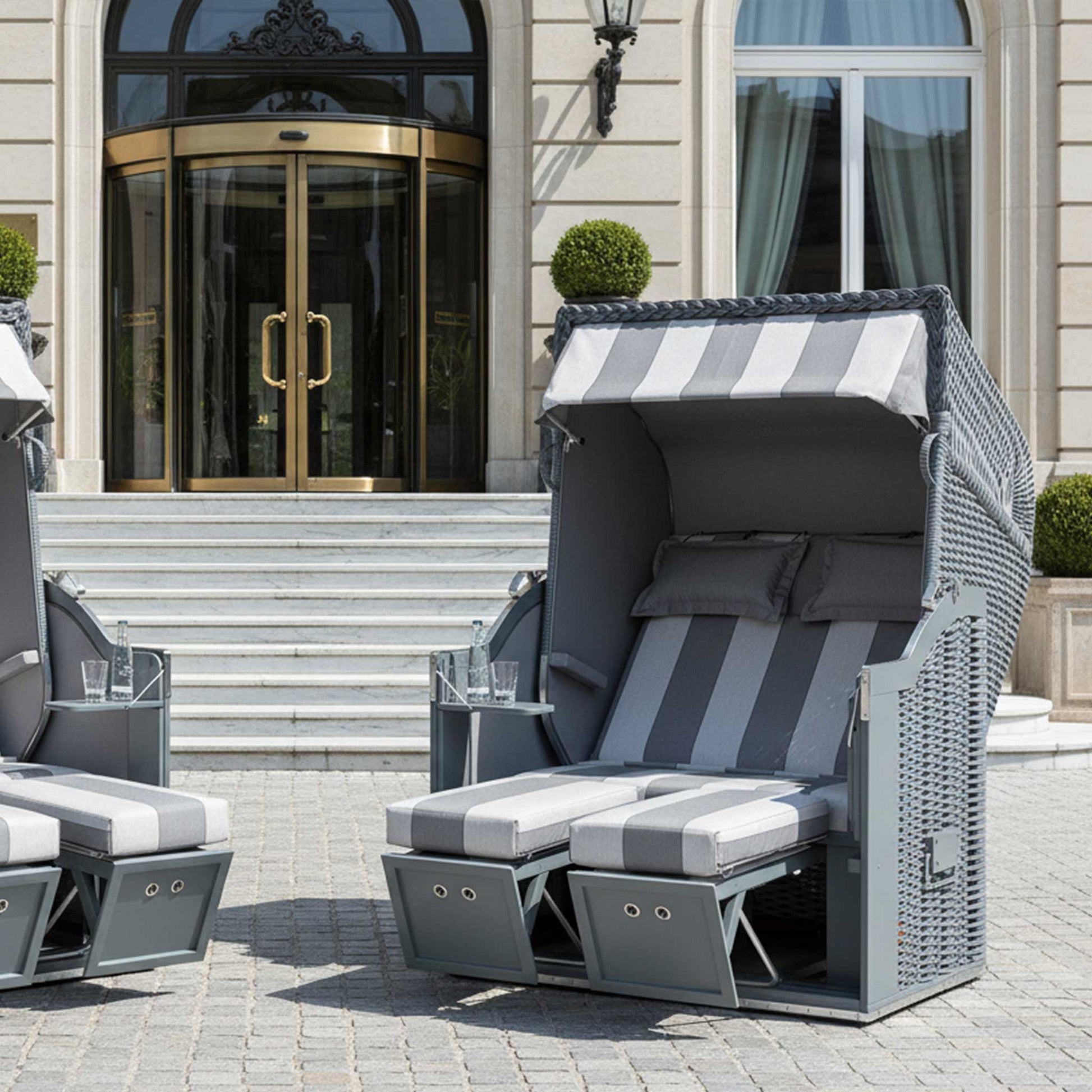 Zwei Strandkorb Sonnenpartner Modelle Classic PE-Geflecht in Onyx Farbe, Holz grau, vor einem eleganten Gebäude.