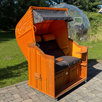 Strandkorb Sonnenpartner Modell Classic PE orange mit schwarz-grauen Kissen im Garten, perfekter Ort zum Entspannen.