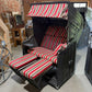 Strandkorb Kiel 255 Plus mit grauem Holz, anthrazitem PVC-Geflecht und gestreiften Kissen in rot und beige.