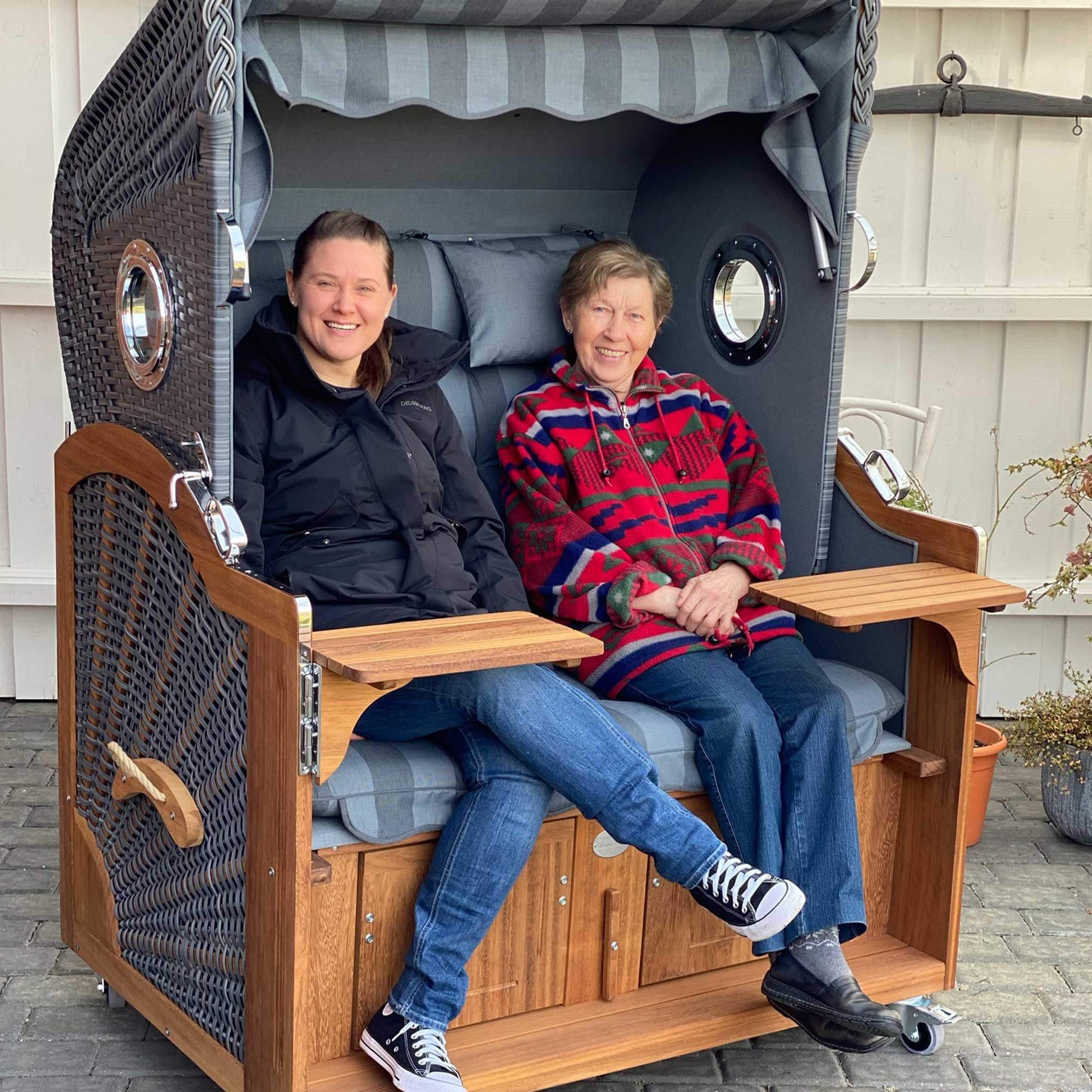 Strandkorb Prieß Modell Meister, zwei Personen sitzen entspannt in einem luxuriösen Strandkorb aus Iroko Holz mit dunkelgrauem Geflecht.