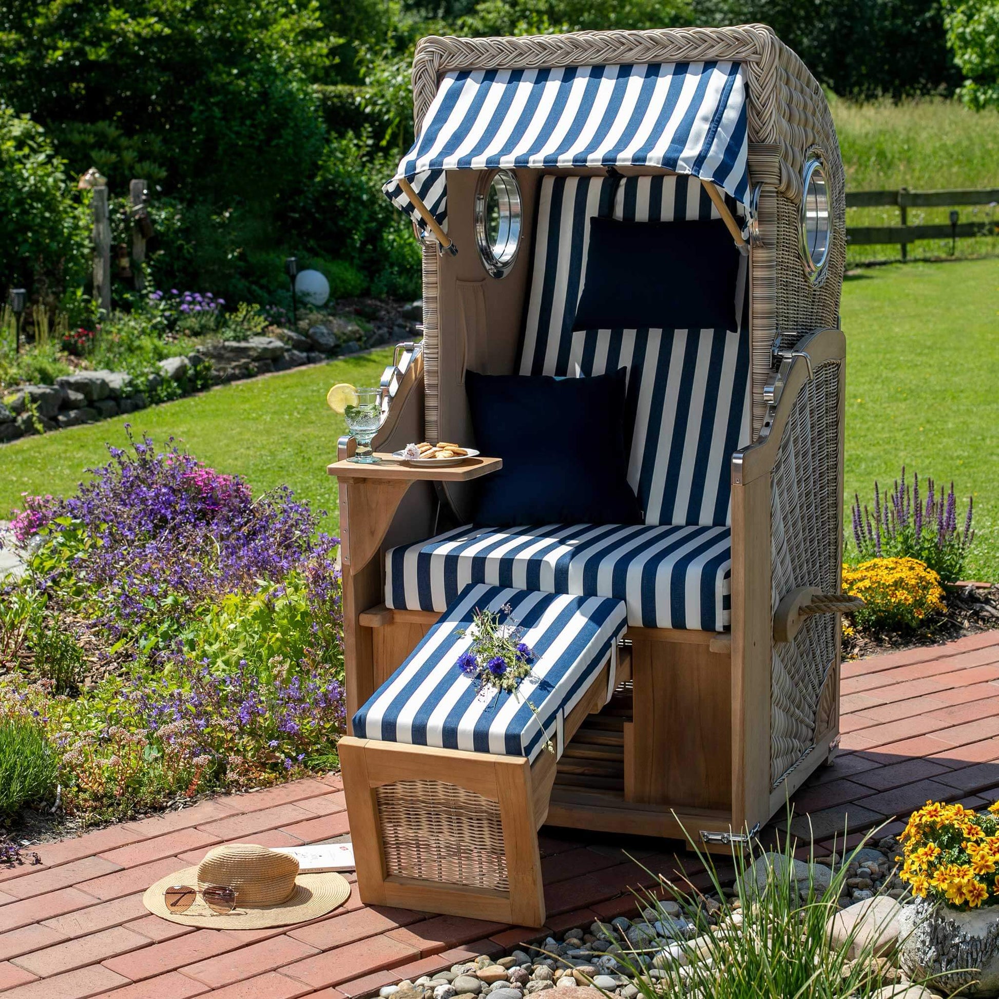 Strandkorb Mr. Deko Düne Bullaugen Teak PE-shell 1-Sitzer mit blau-weißem Dessin 518 im sommerlichen Garten auf Terrasse