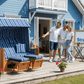 XL Strandkorb SunnySmart Rustikal 250 Plus auf Holzterrasse vor blauem Haus mit Paaren, blau weiße Polster, Halbliege XL-Sitzer